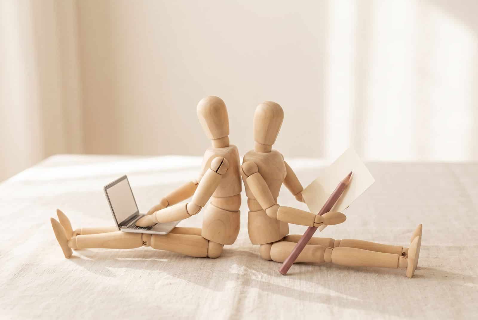 Two wooden mannequins back to back, one on a laptop, one with paper and pencil